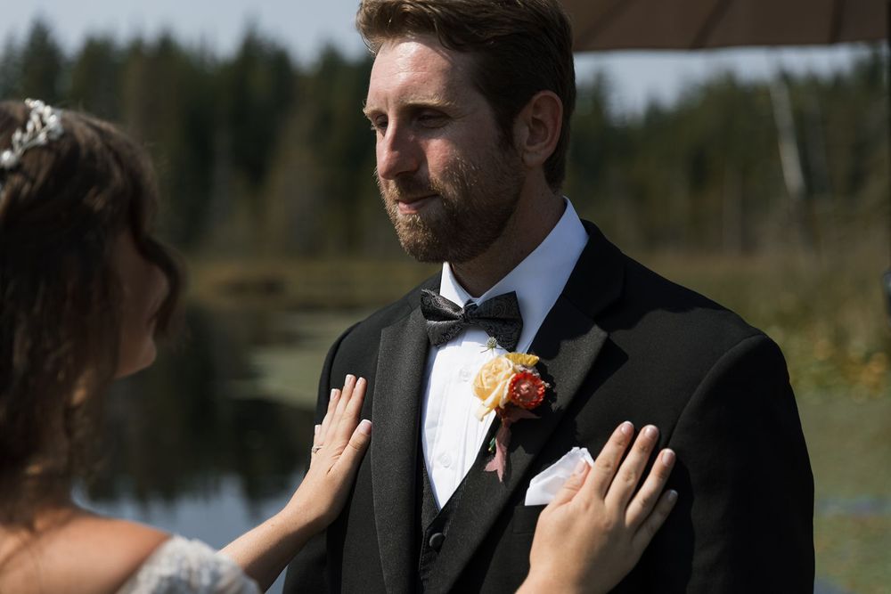 Emotional groom in black tuxedo with yellow boutonniere during a first look near a scenic lake.