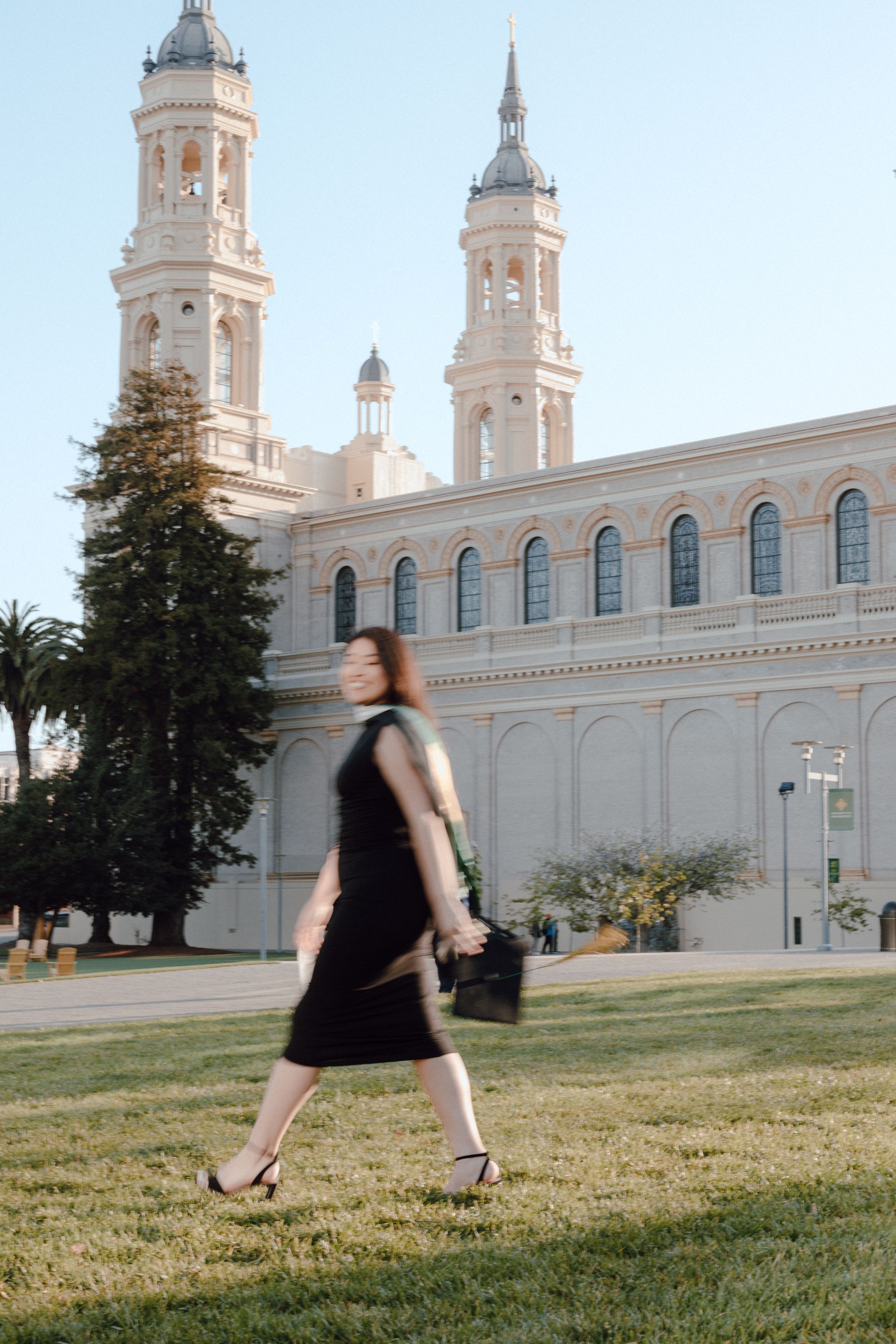 Graduate holds cap and walks across lawn of St. Ignatius Church at the University of San Francisco. By Bay Area Graduation Photographer.