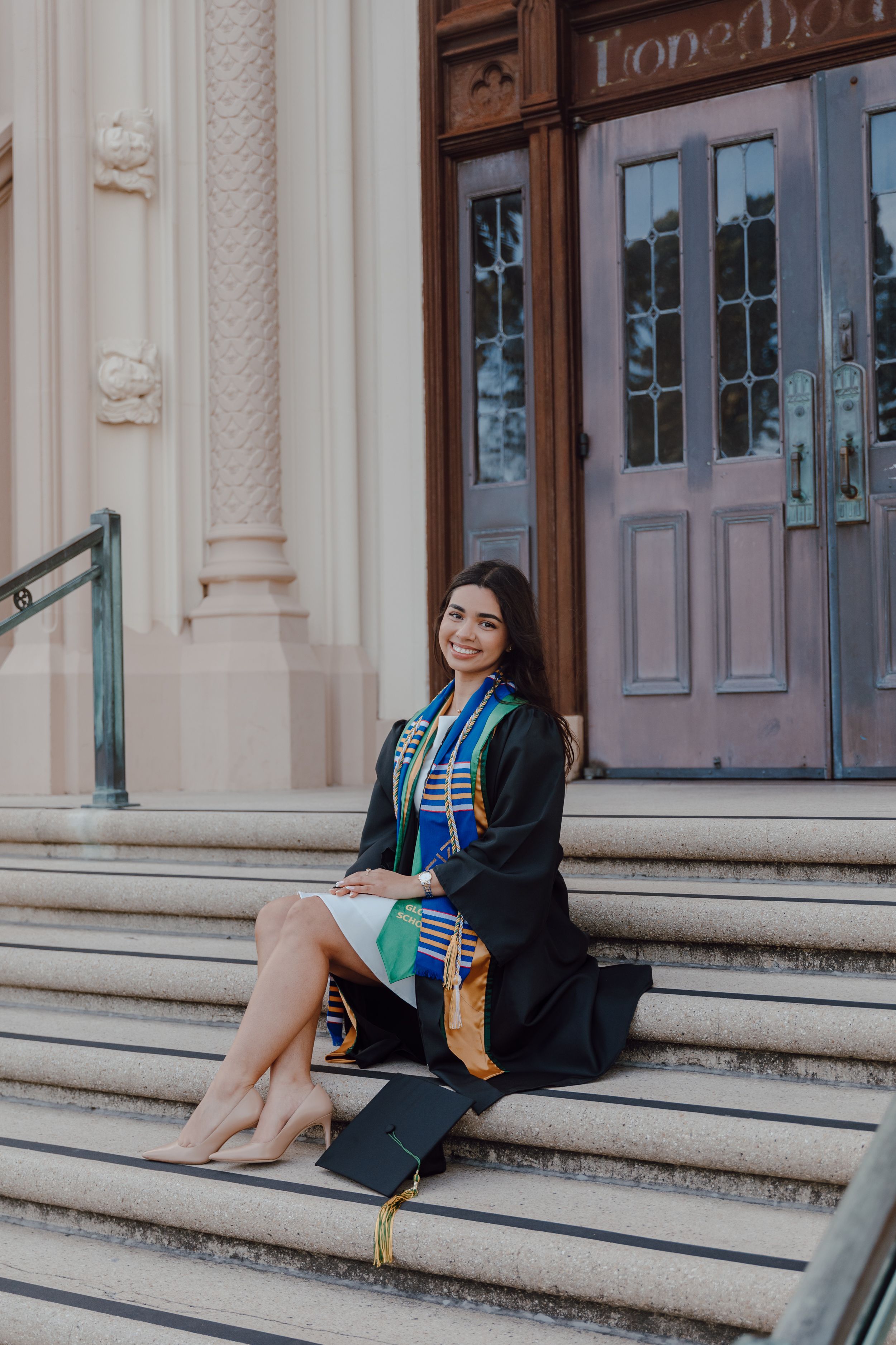 Graduate sits on steps in front of Lone Mountain at the University of San Francisco. Cap sits beside her Bay Area Graduation Photographer.