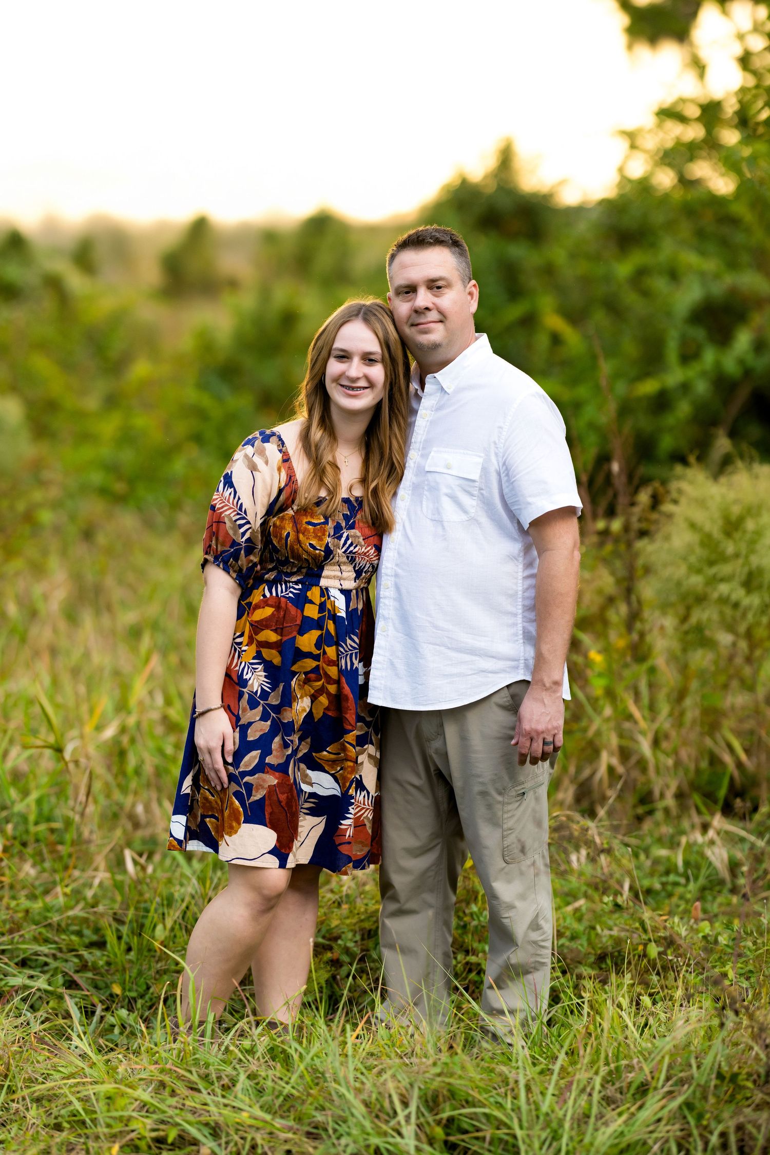 A couple stands together in a grassy field during golden hour sunset, wearing casual summer attire.