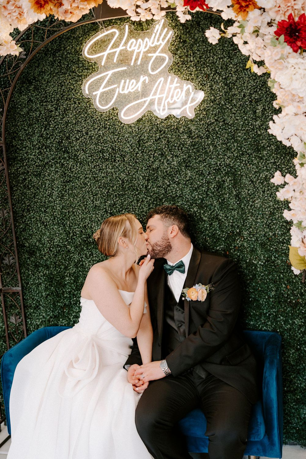 Romantic wedding couple sharing a kiss on a blue couch against a floral greenery backdrop with neon signage.