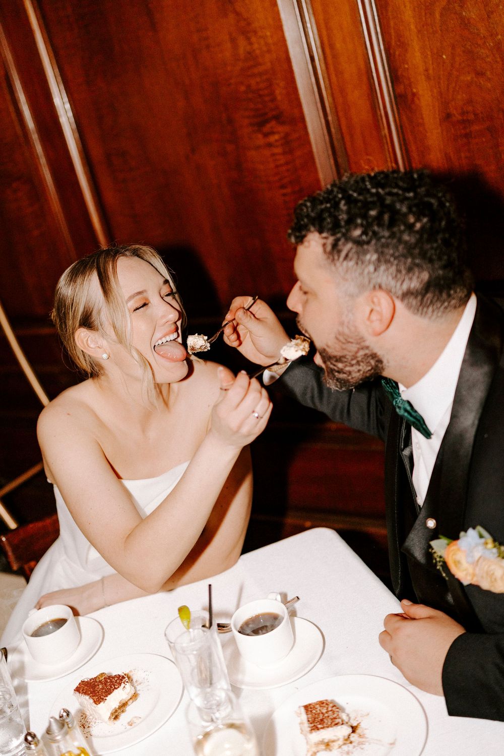A couple shares a joyful moment feeding each other dessert at an elegant dinner table with warm lighting.