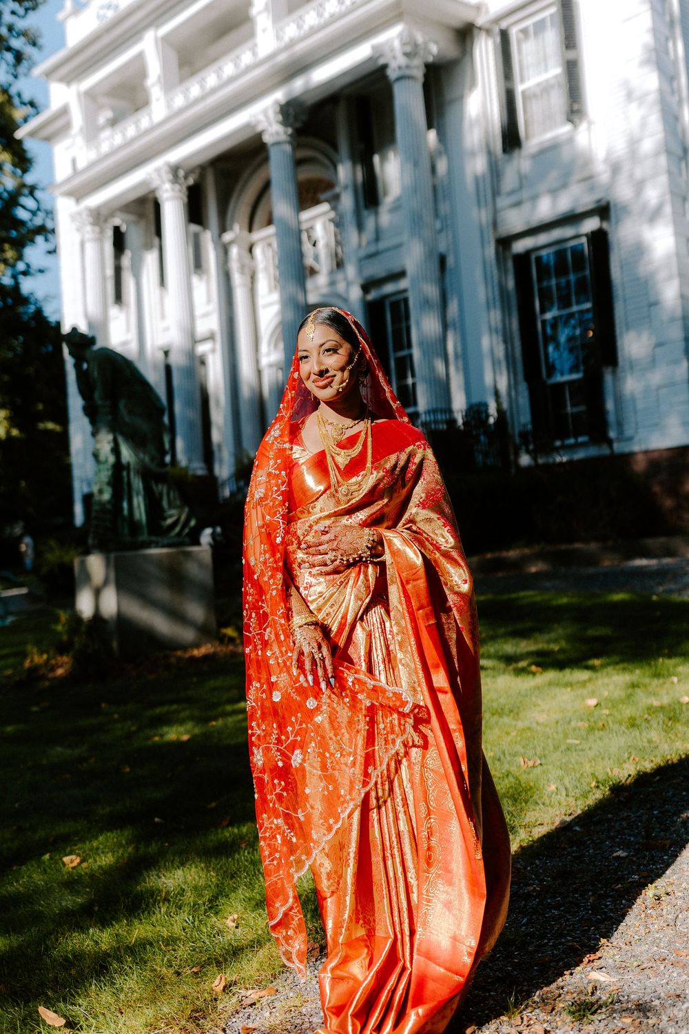 A bride in a vibrant red and gold traditional Indian saree with dupatta stands smiling outdoors near a white colonial building.