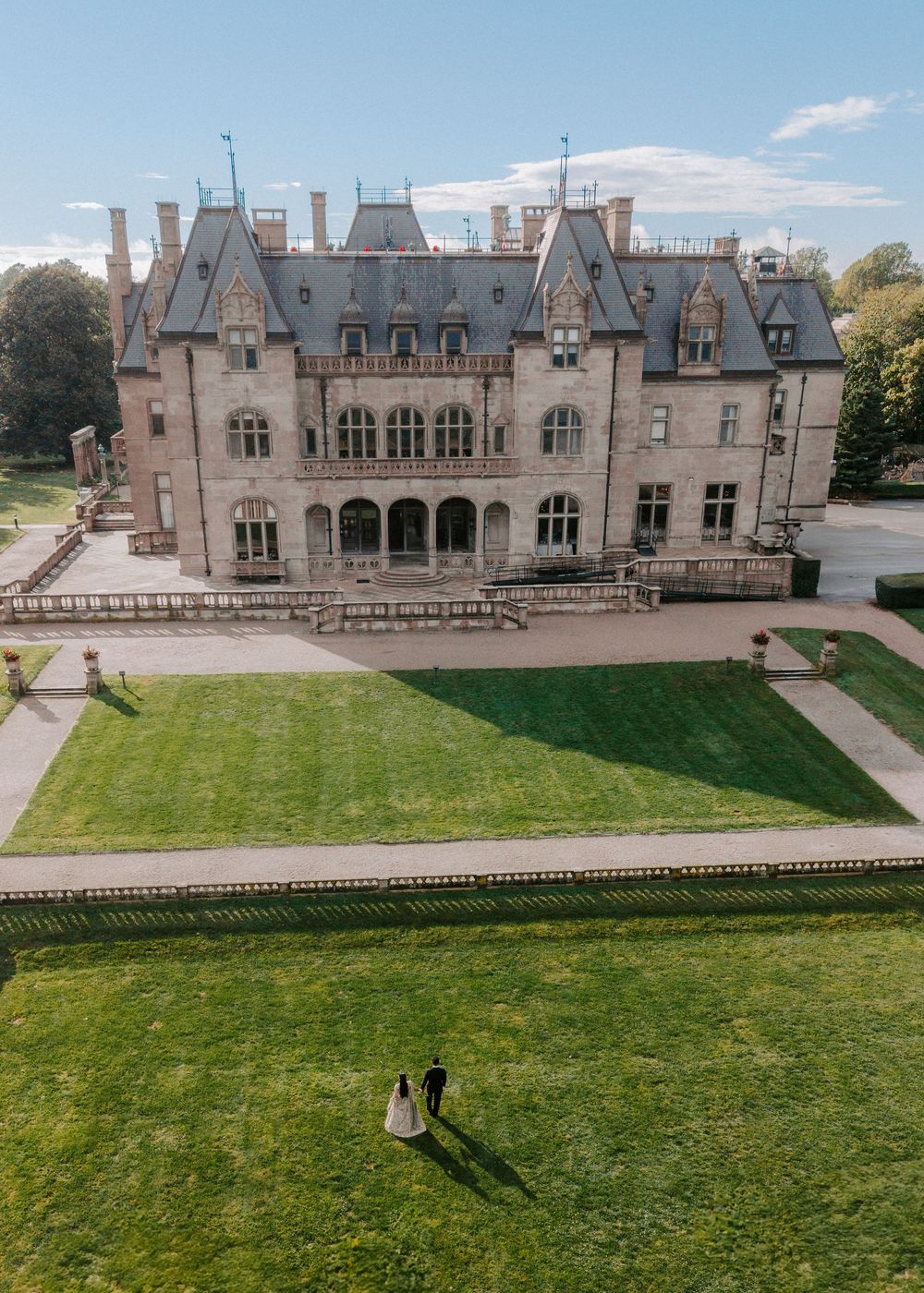 Aerial view of a bride and groom standing on lush green lawn before a grand historic mansion on a sunny day.
