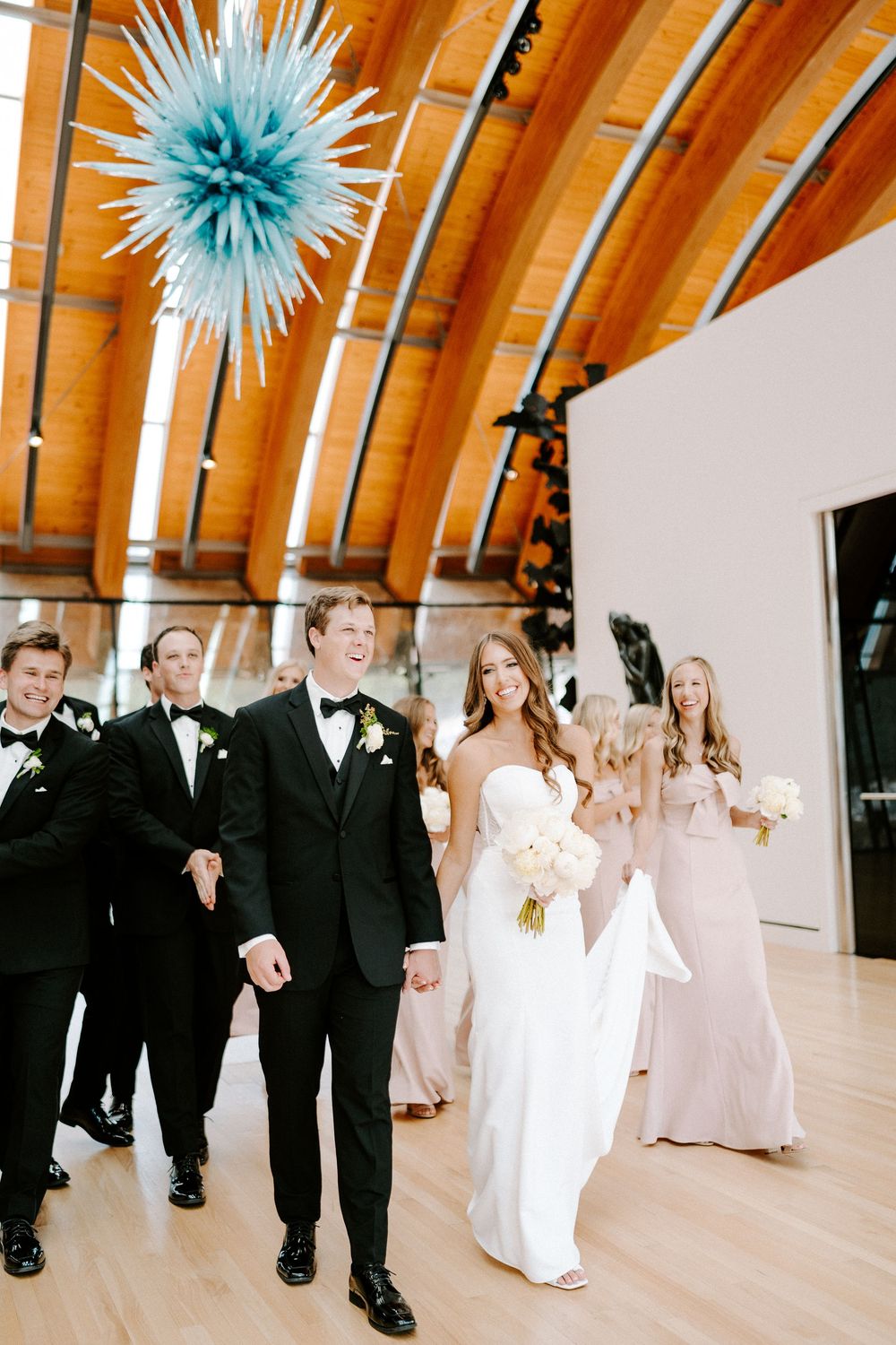 Wedding party in formal attire poses beneath blue decorative ceiling ornaments in an elegant venue.