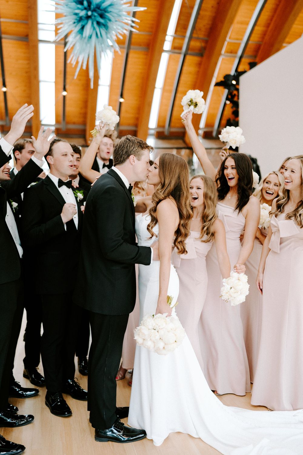 Wedding celebration with bridal party in white dresses and black tuxedos dancing in a bright venue.