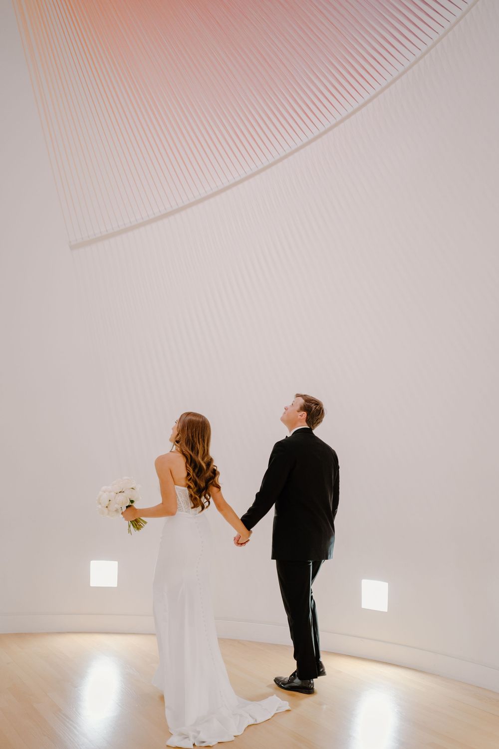 A couple in wedding attire holds hands while standing in a minimalist white curved room with floor lighting.
