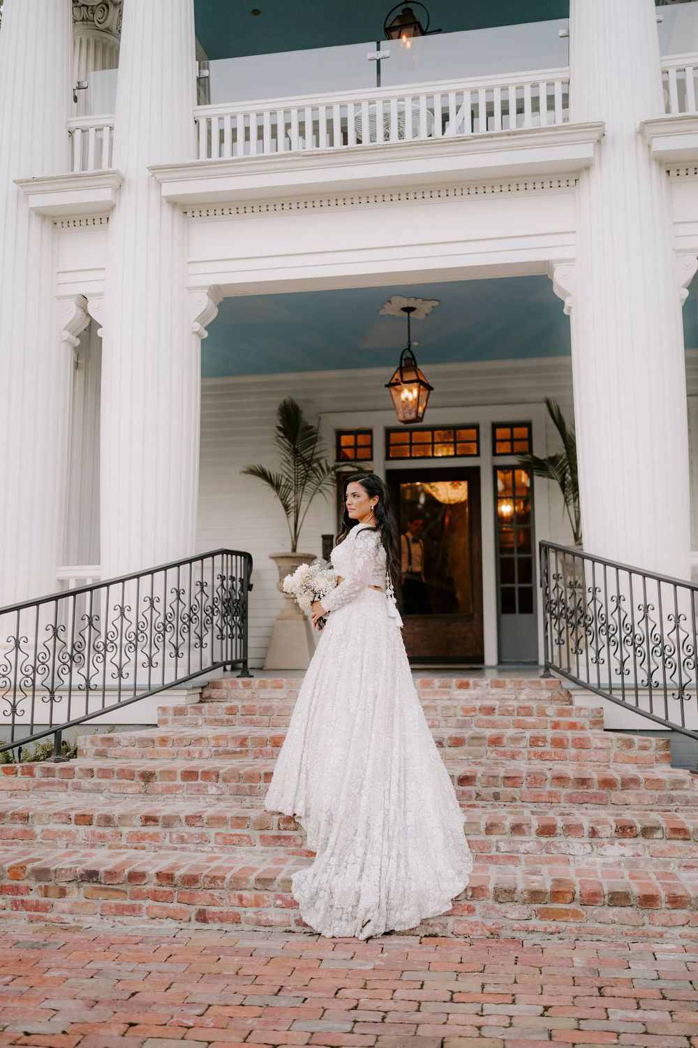 A bride in a vintage-style lace wedding dress stands on brick steps of an elegant white columned mansion entrance.