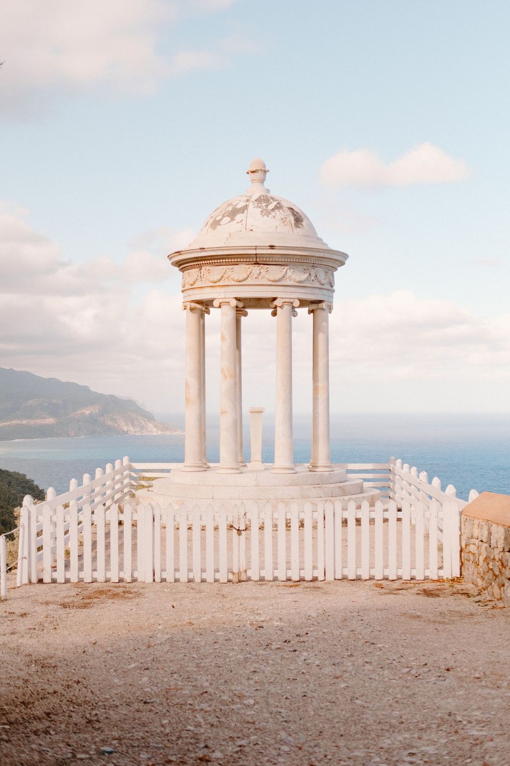 White marble gazebo with columns and picket fence overlooks a serene blue sea under a partly cloudy sky.