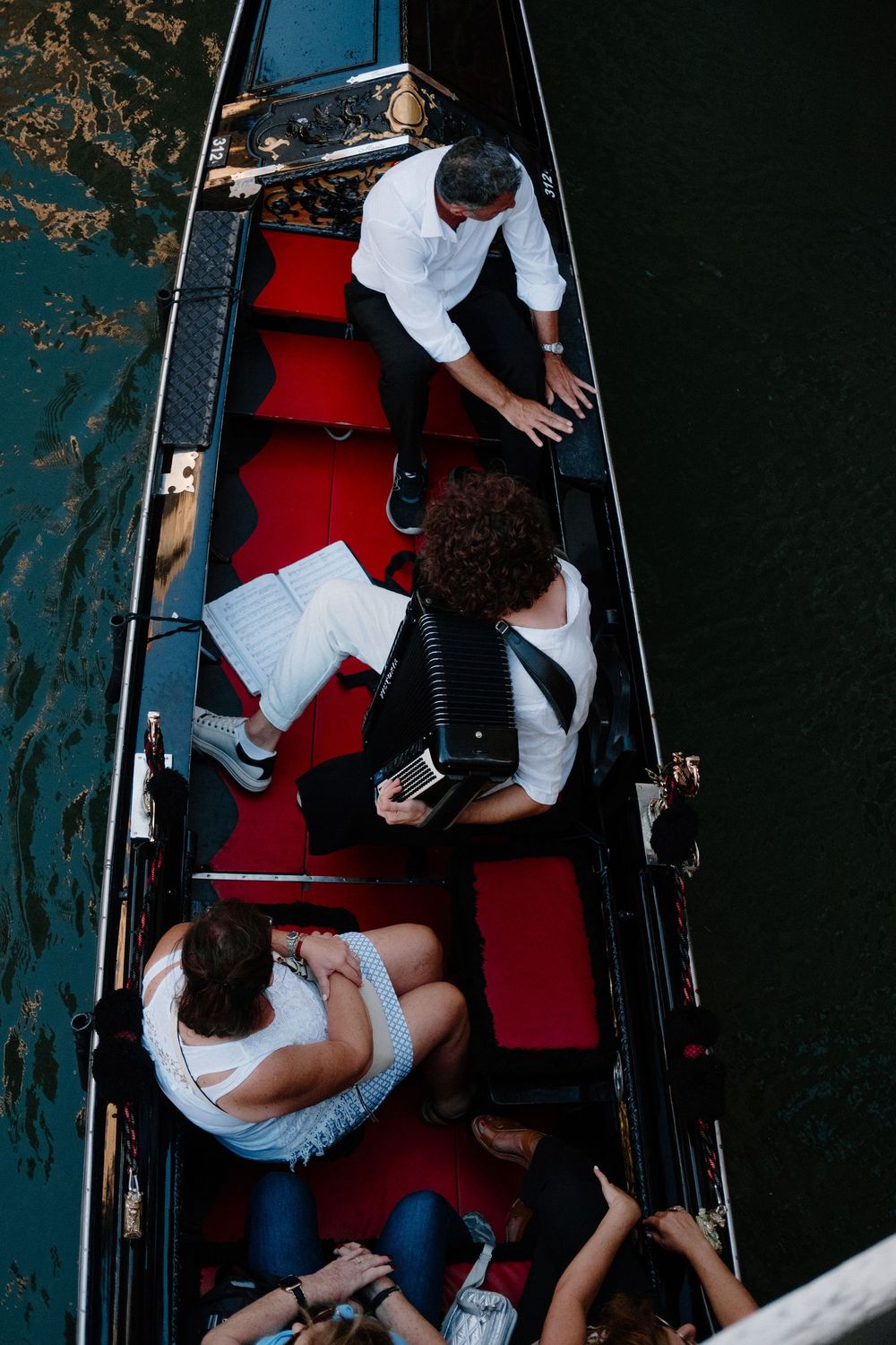Aerial view of tourists riding a traditional red-seated gondola on a Venice canal with a gondolier in white shirt.