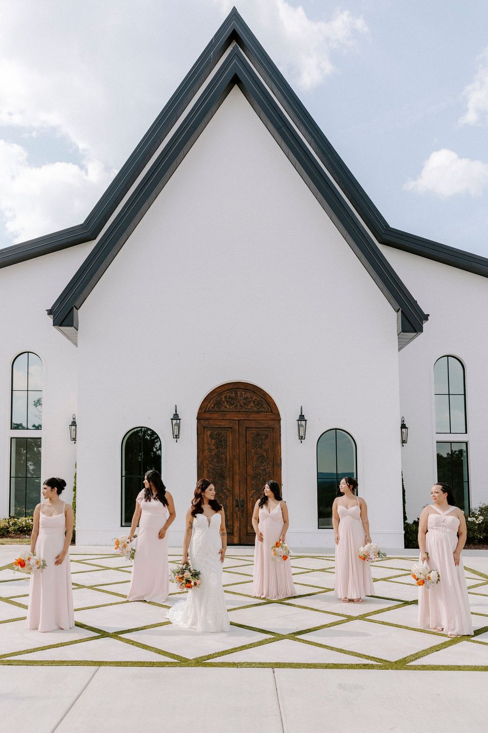 Bride and bridesmaids in blush gowns pose outside a white chapel with a grand wooden door.