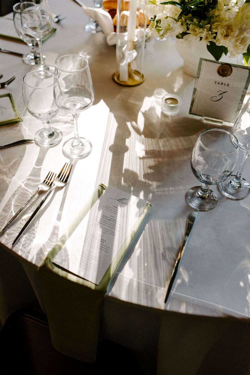 Elegant wedding reception table setting with white linens, crystal glasses, silverware, and floral centerpiece in natural light.