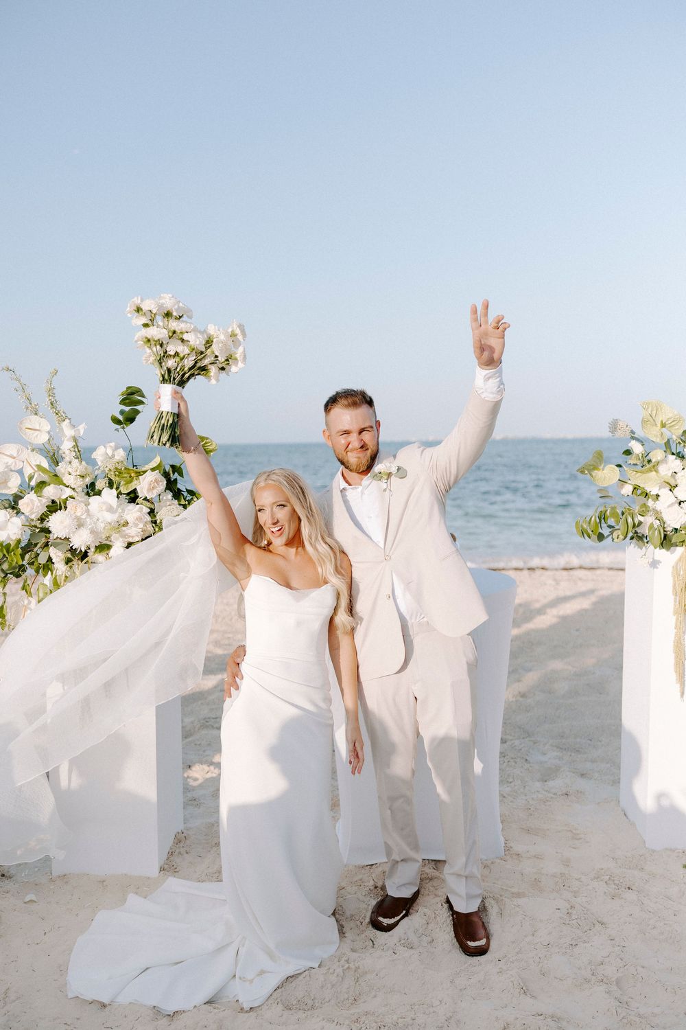 Wedding couple celebrating with raised arms on a beach with white floral decorations.