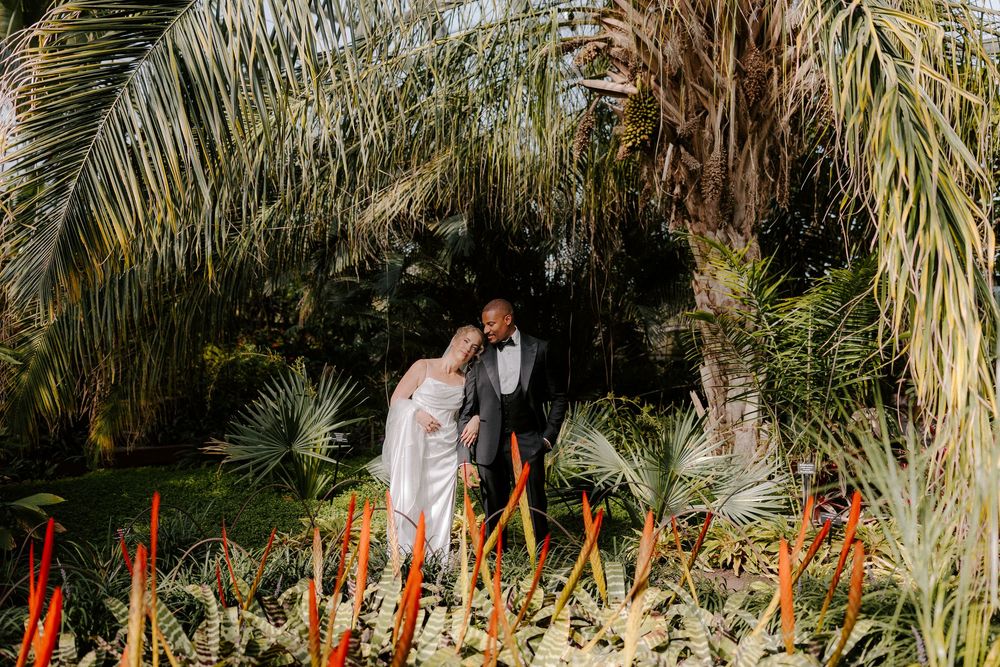 Bride and groom stand among tropical palms and red plants in a lush garden wedding portrait.