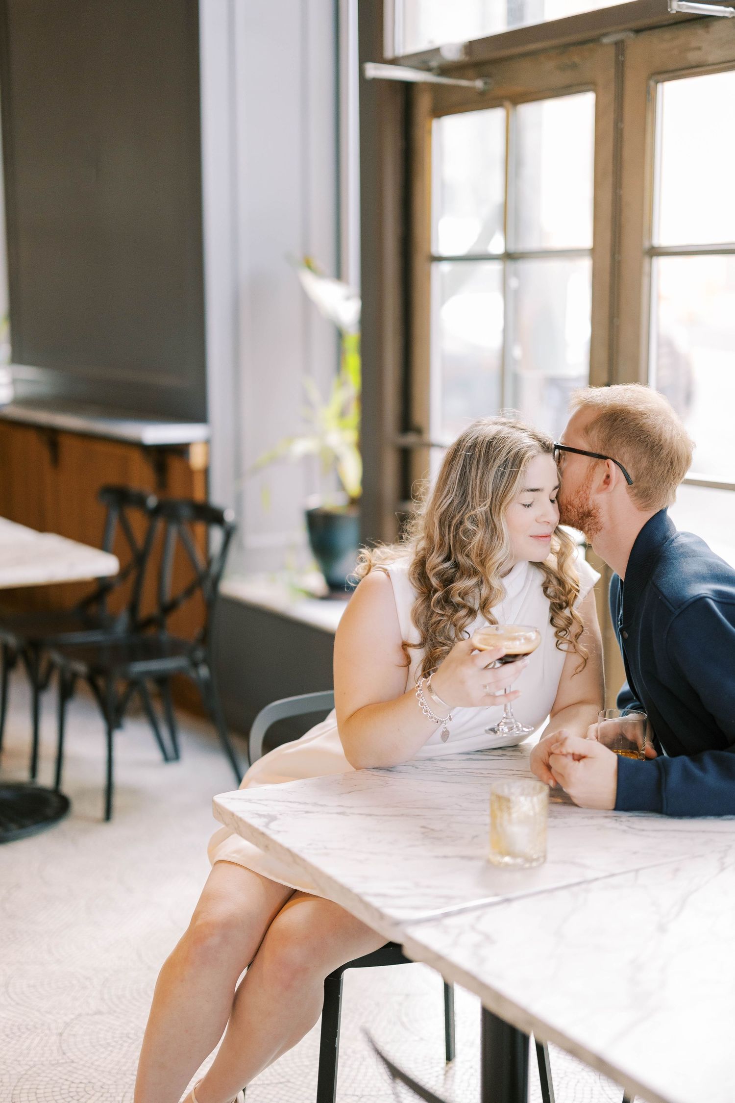 Romantic couple shares intimate moments at a sunlit table by large windows in a bright restaurant setting.