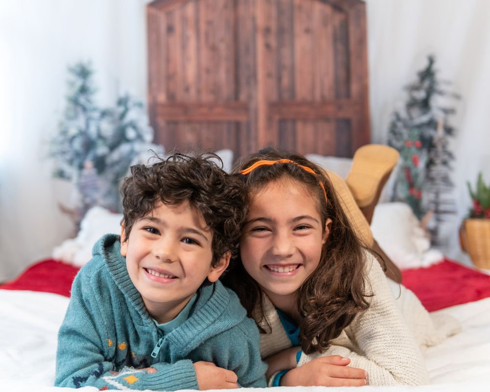 Two siblings pose together on white bedding with wooden decor in background.