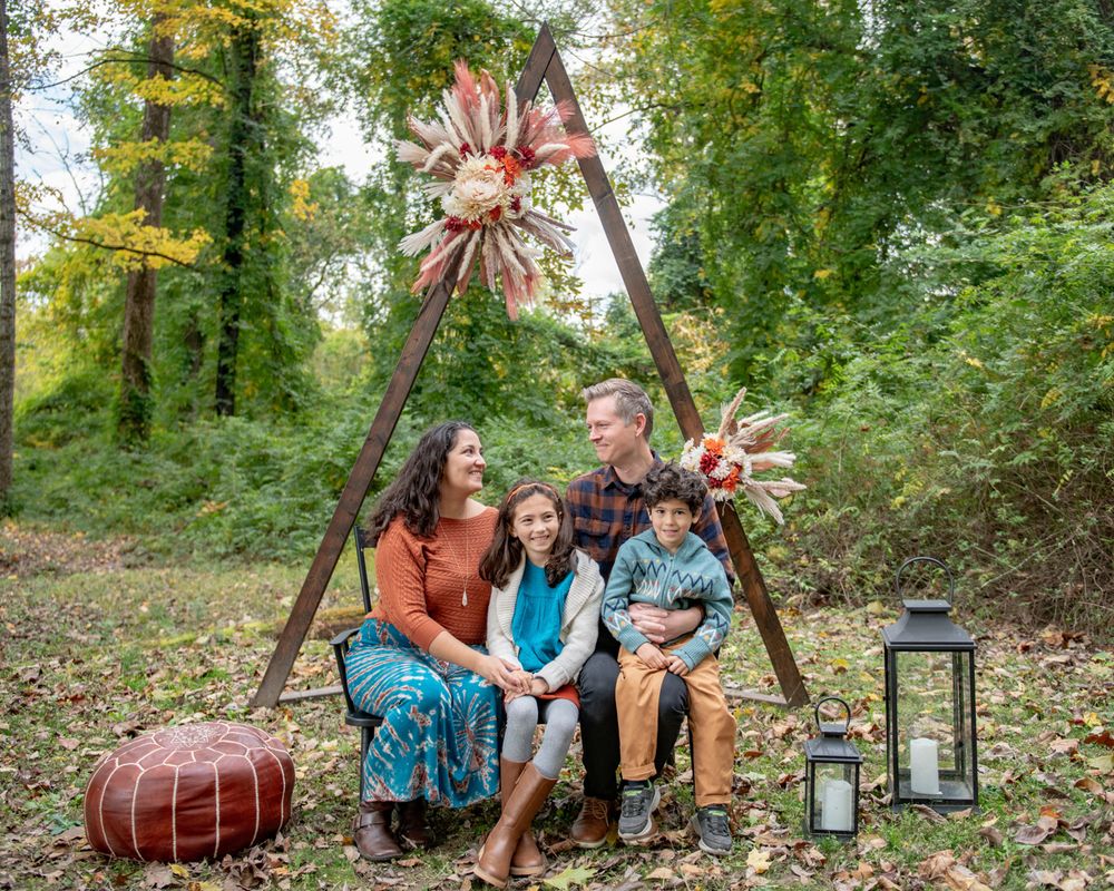 Family sits on triangular wooden swing decorated with floral arrangements in forest setting.