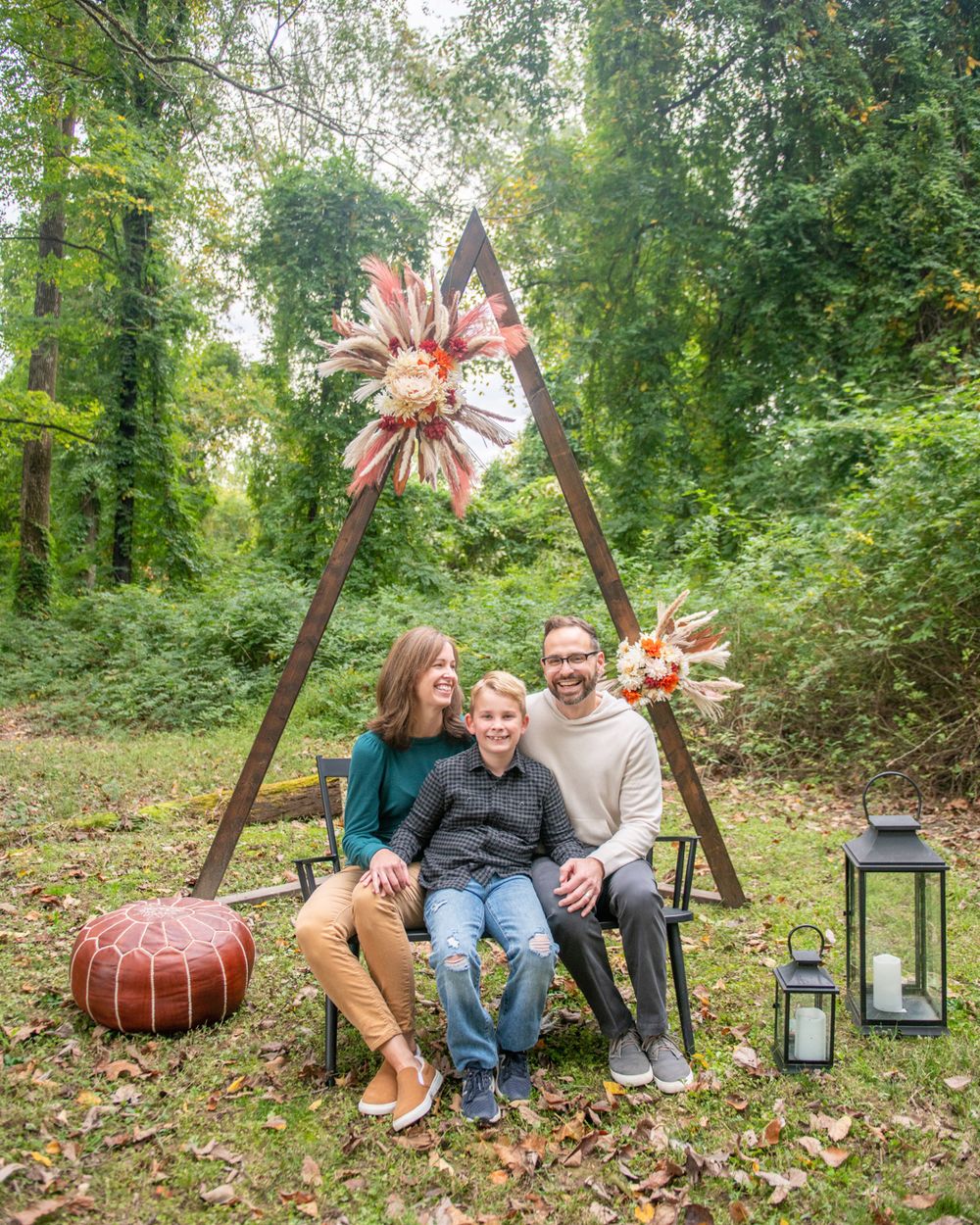Family poses on wooden swing frame with decorative flowers in wooded outdoor setting.