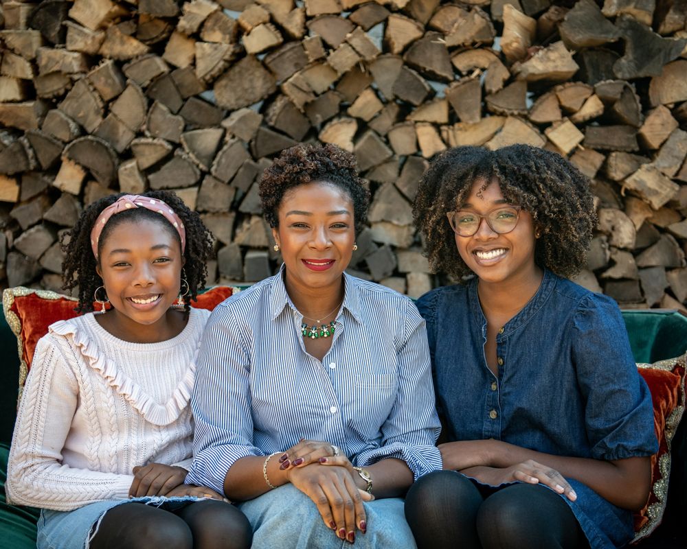 Three friends sitting on outdoor couch against stacked firewood background.