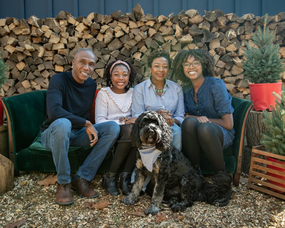 Family of four with dog sitting on green couch against woodpile backdrop.