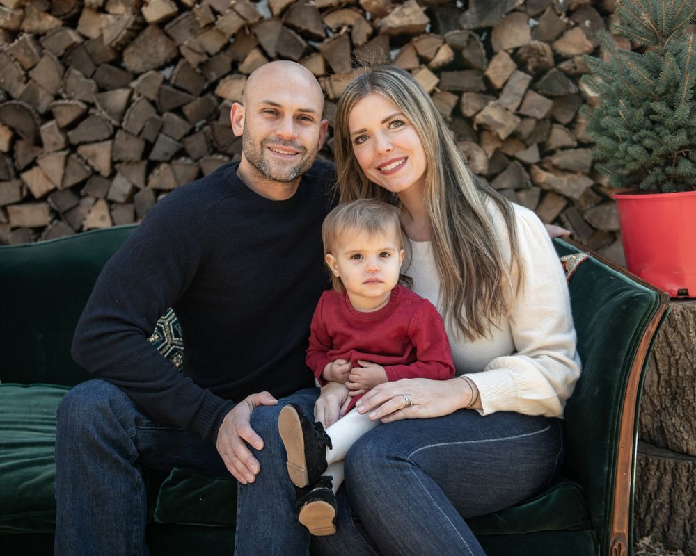 Family of three poses together on vintage green couch against stacked firewood.