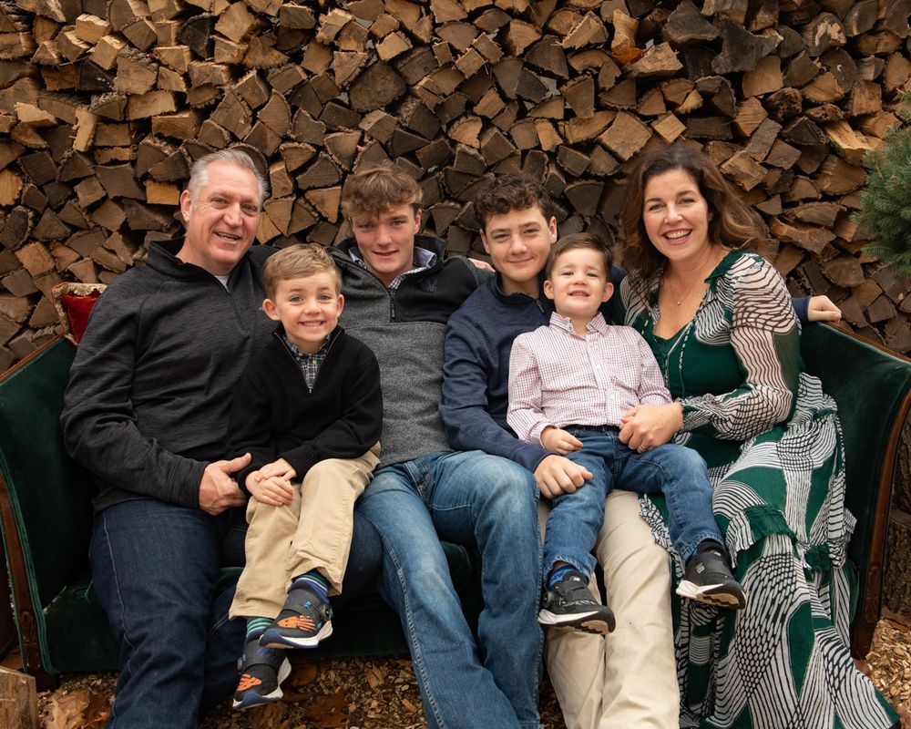 A family group poses together on a bench against a rustic woodpile backdrop.