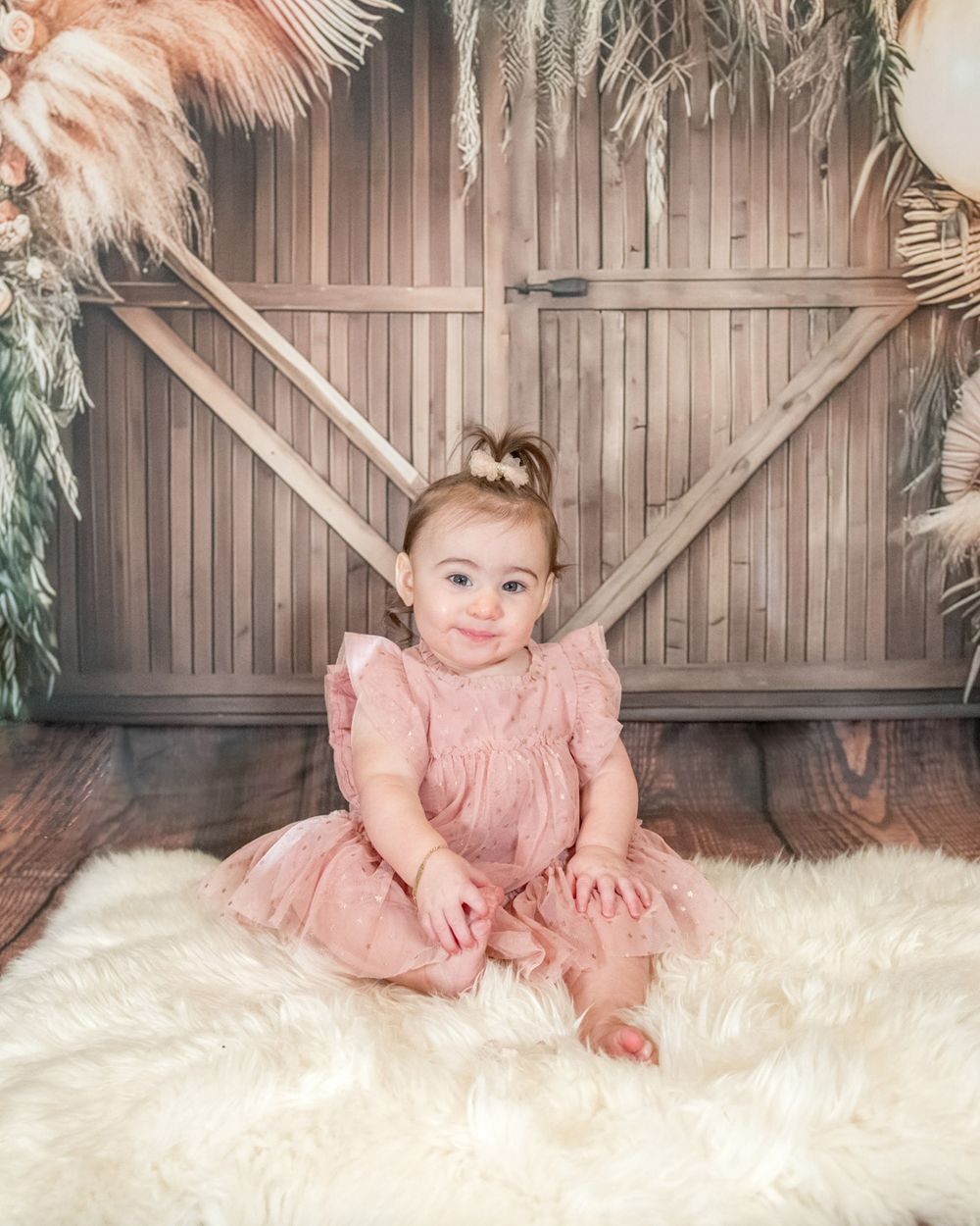 Baby in pink dress sits on white fur rug against rustic barn door backdrop.