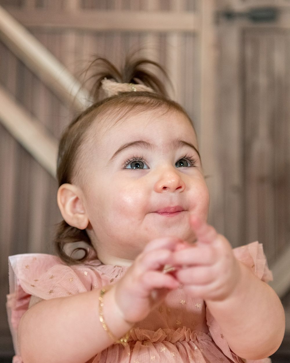 Baby wearing pink lace dress smiles while clapping hands against rustic barn door backdrop.
