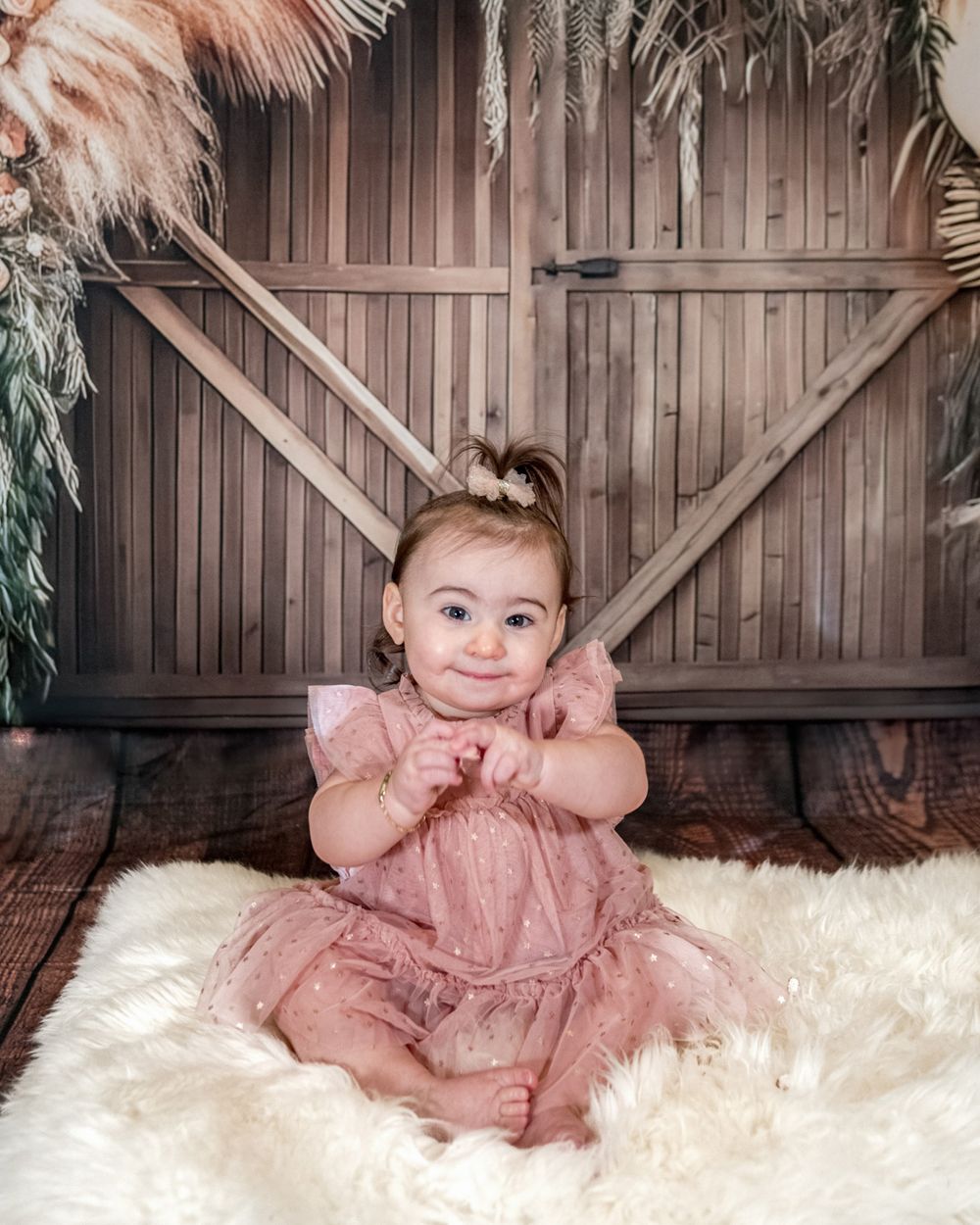 Baby in pink dress smiles while sitting on white fur against wooden backdrop with decorative foliage.