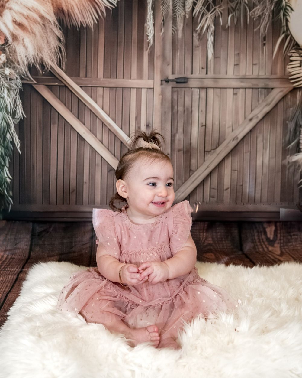 Toddler in pink dress smiles while sitting on white fur against rustic backdrop.
