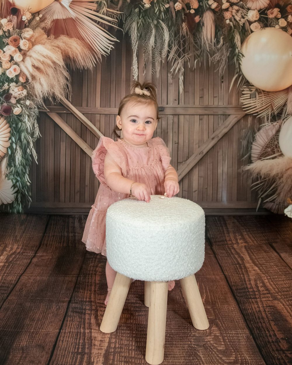 Baby in pink dress stands beside white ottoman against decorated wooden backdrop.
