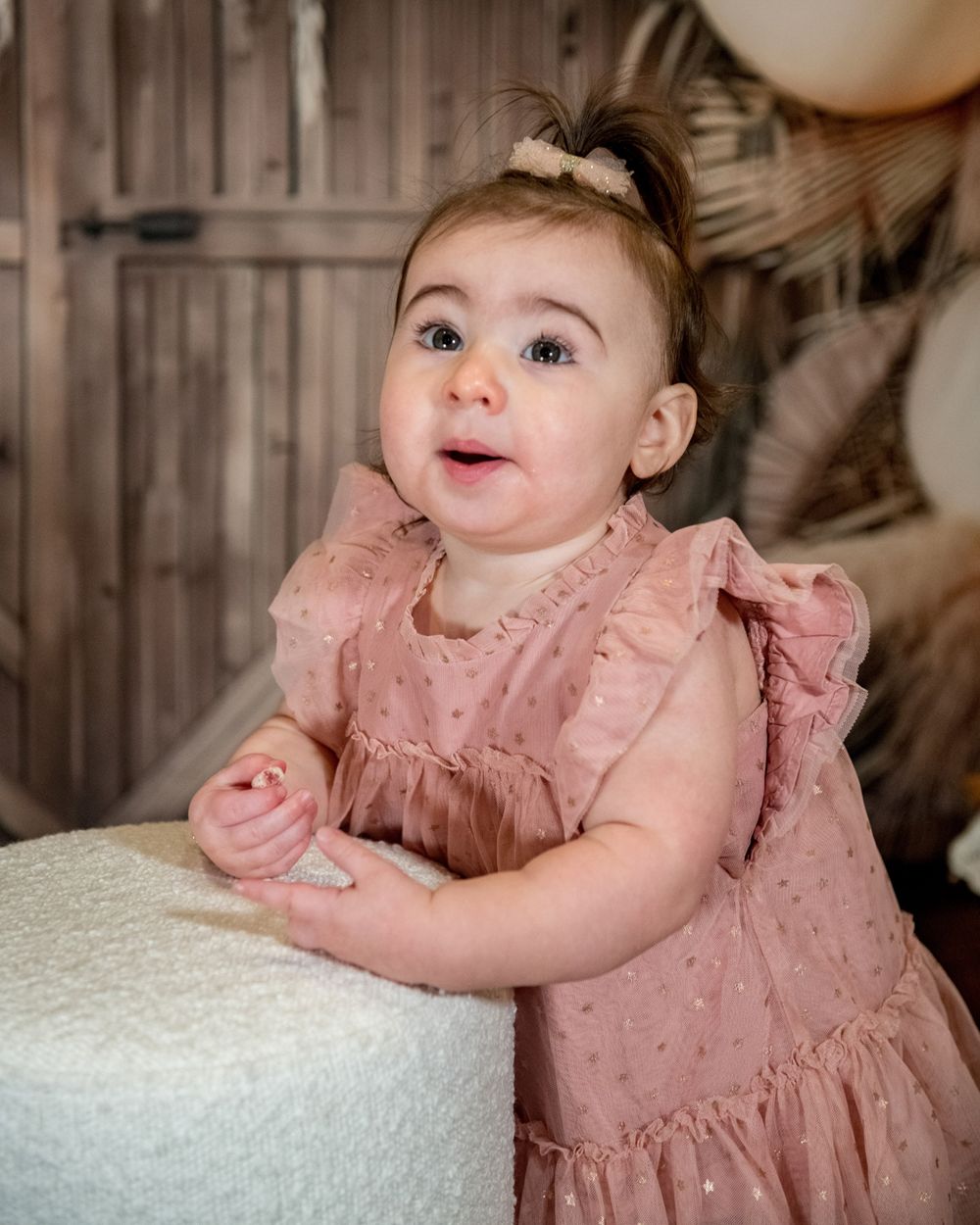 Baby in pink ruffled dress leans on white ottoman with decorated backdrop.