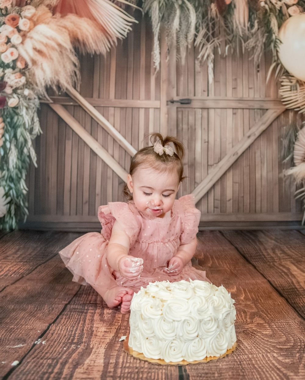 Baby in pink dress reaches towards white frosted cake on wooden floor with decorated backdrop.