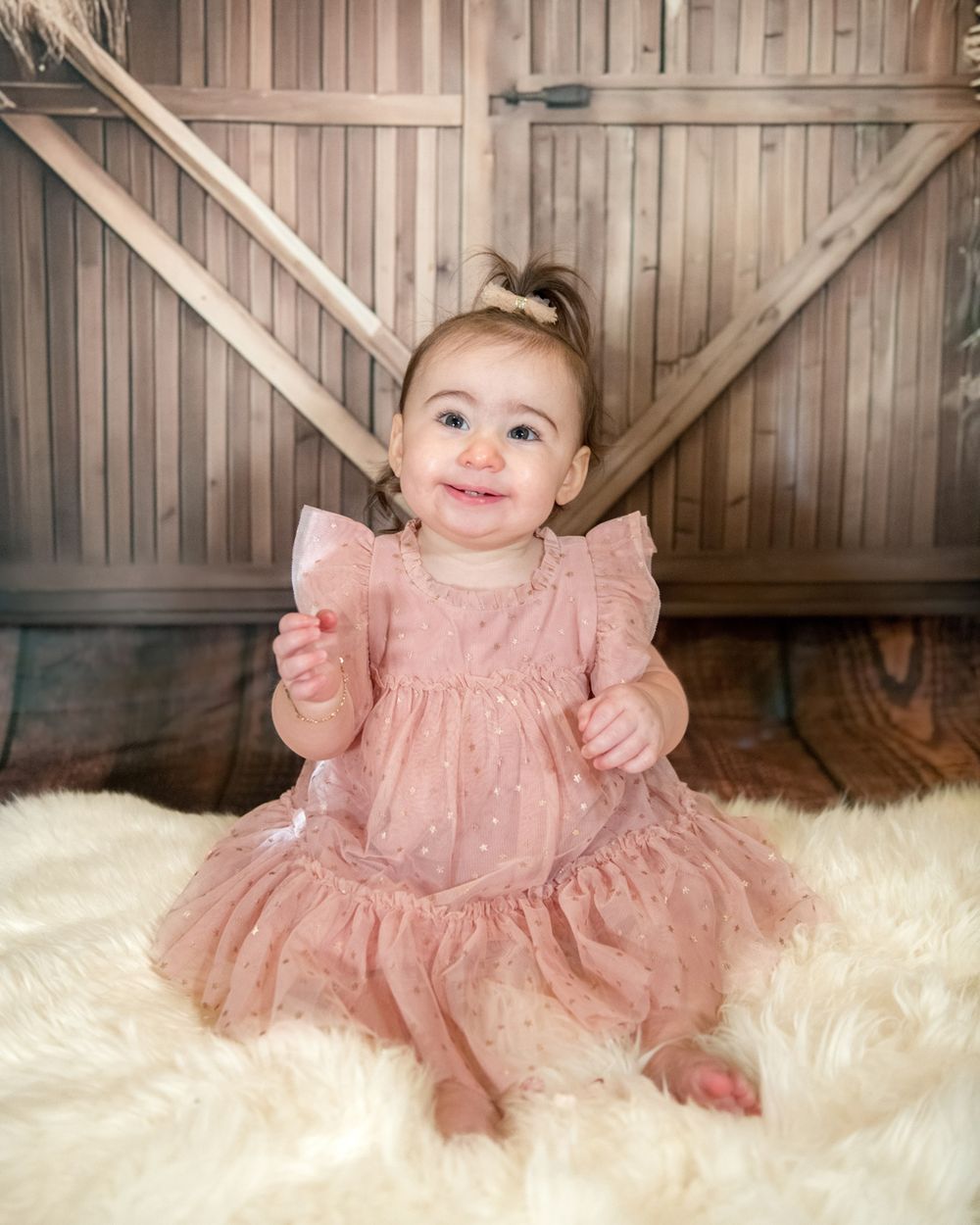 Baby in ruffled pink dress sits on white fur rug against wooden barn door background.