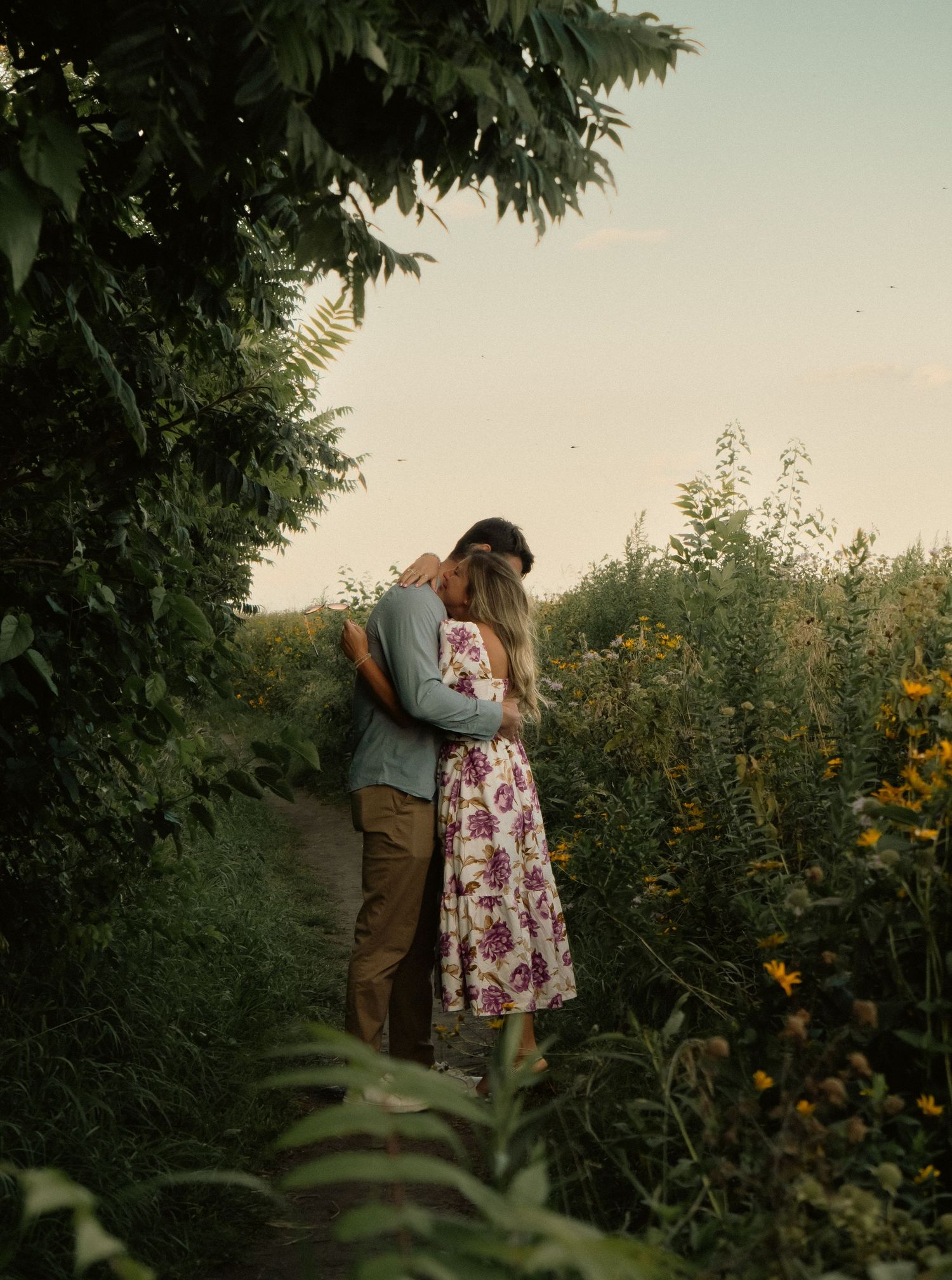 A romantic couple embraces in a garden with wildflowers and trees at sunset.