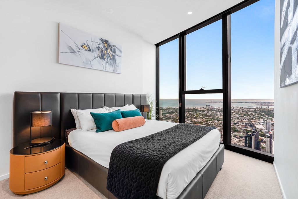 Modern bedroom with black headboard, orange nightstands and floor-to-ceiling windows overlooking a city skyline.