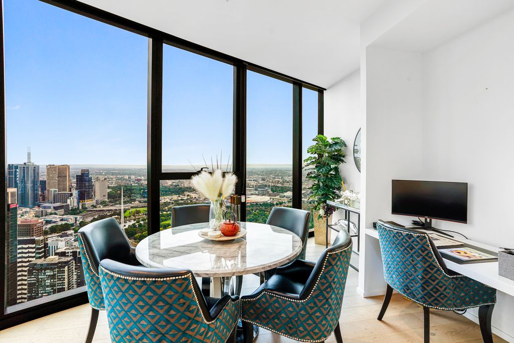 Elegant dining area with teal chairs and round table next to large windows showcasing city views.