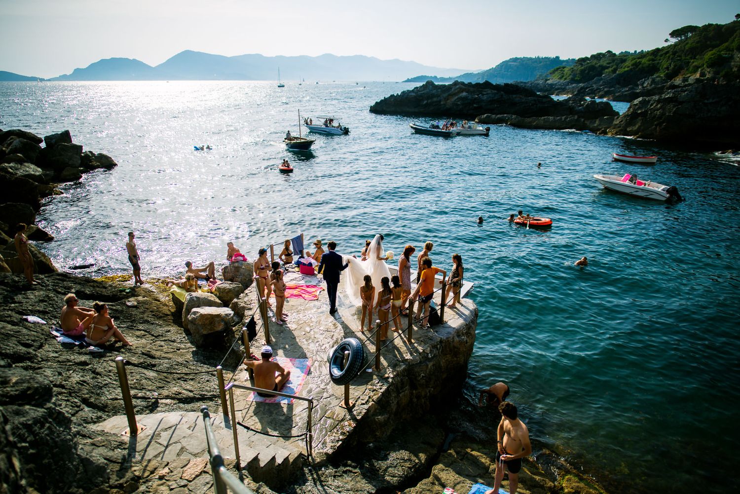 Crowd gathered on rocky coastline with boats in Mediterranean-style bay during sunny day.