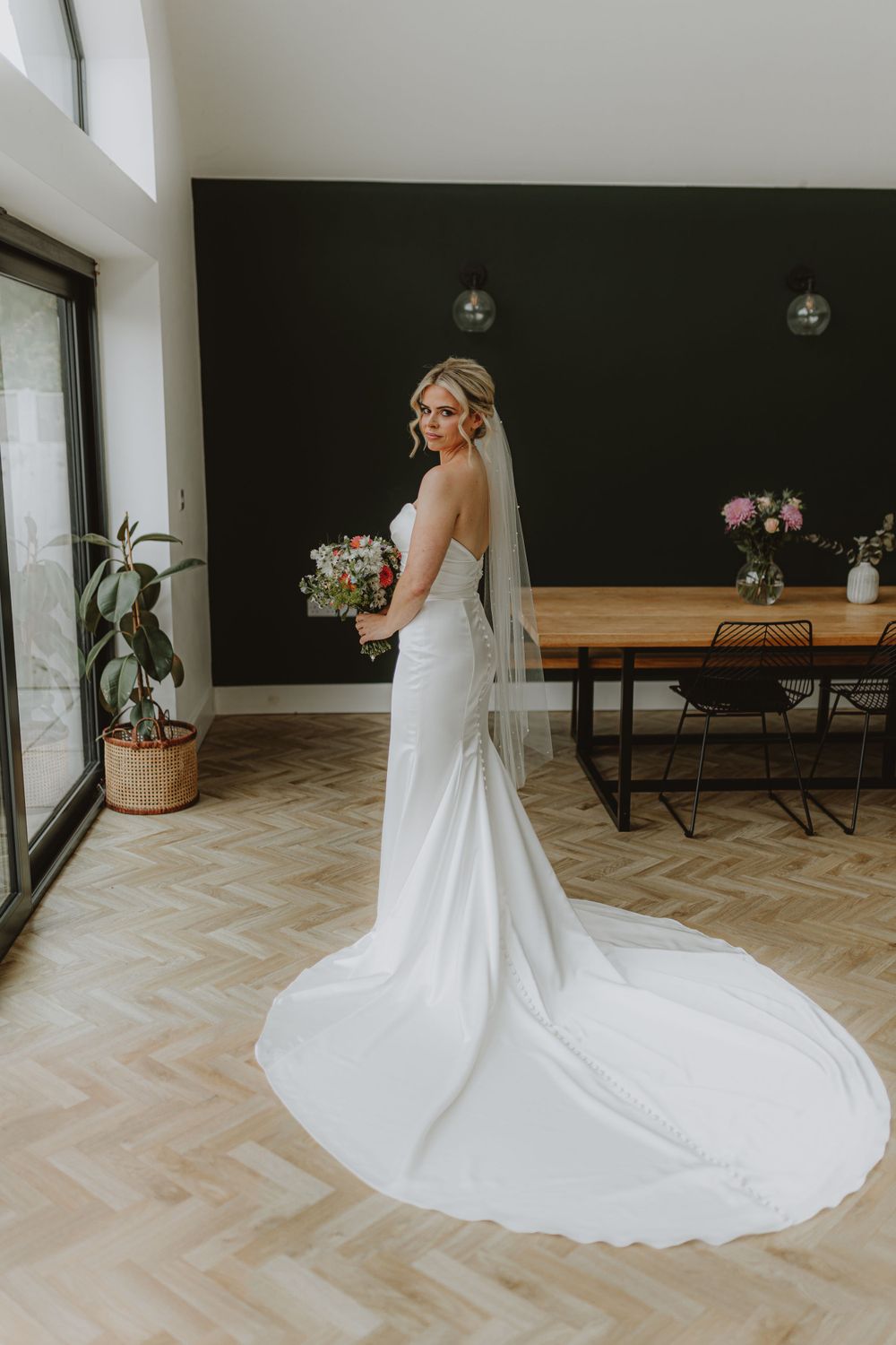 A flowing white wedding dress with a long train displayed in a bright interior space with herringbone floors.