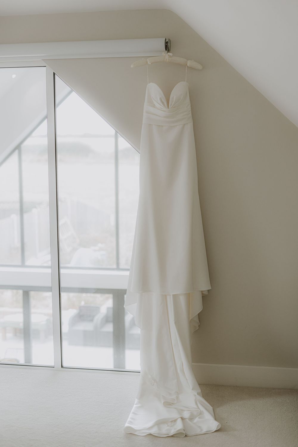 A white wedding gown hanging by a bright window in a minimalist room setting.