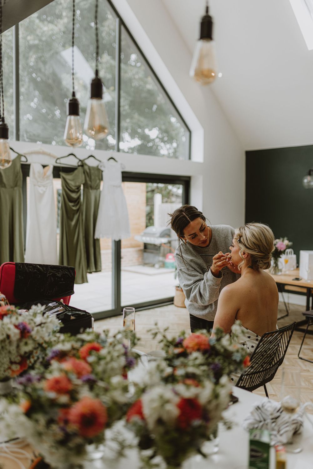 Wedding preparations in a bright modern room with hanging dresses and floral arrangements.