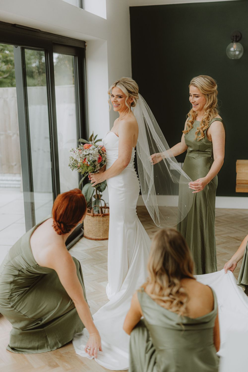Bridesmaids in olive green dresses assisting with the bride's dress and veil.