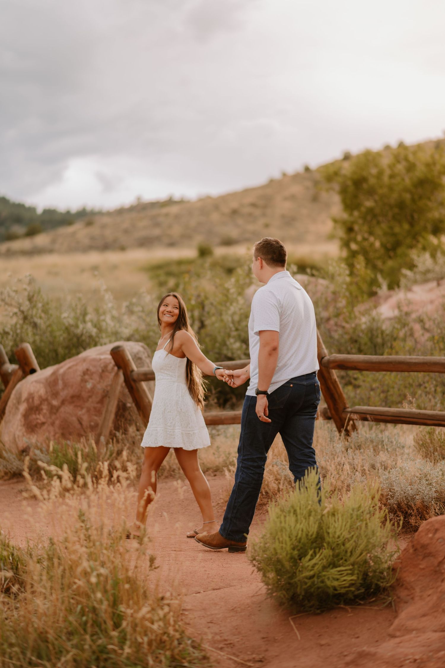 A woman in a white dress holding hands with a man in a white shirt, walking on a trail with rocky and grassy surrounding