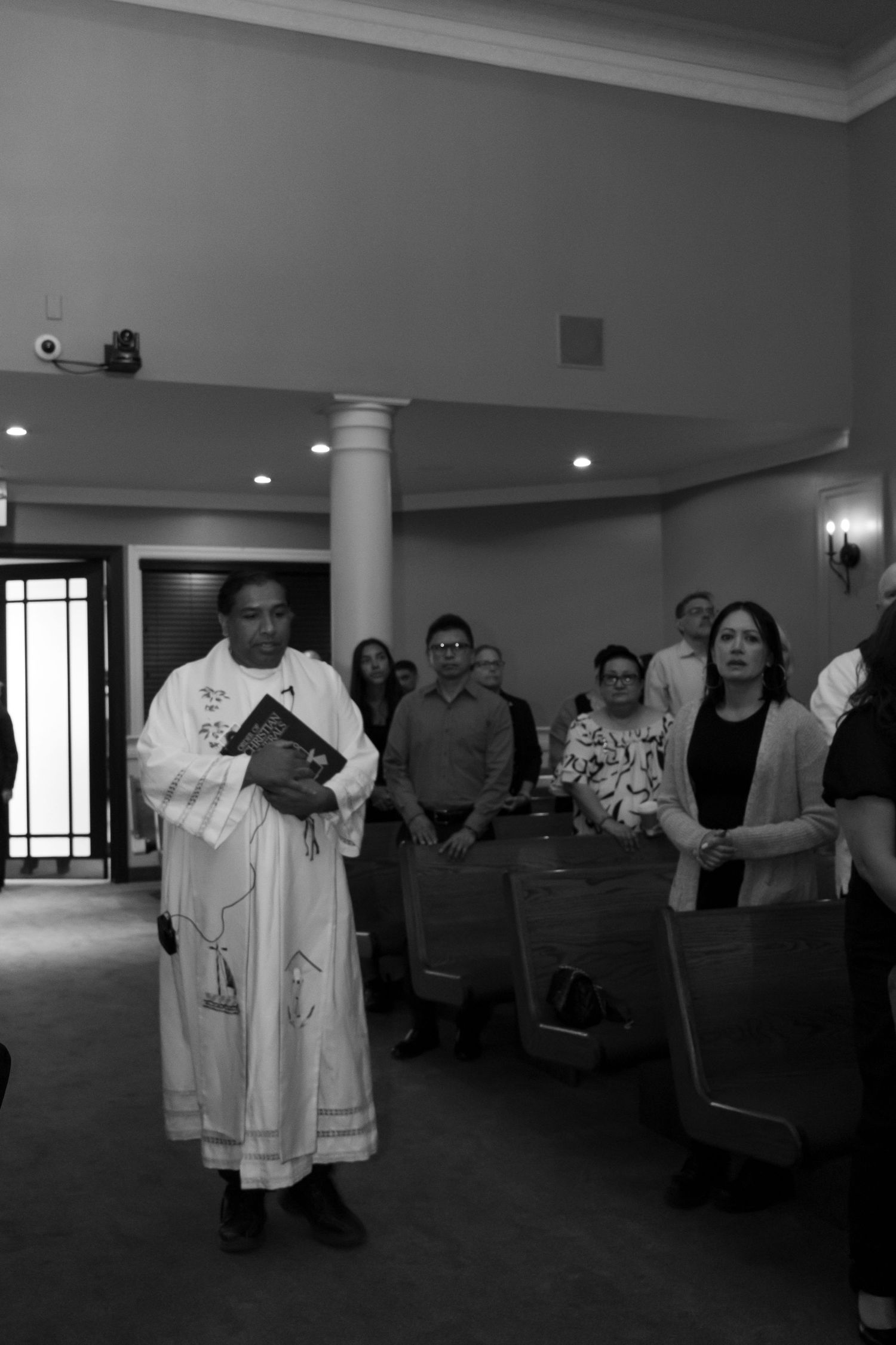 Black and white photo of a priest walking down an aisle surrounded by people