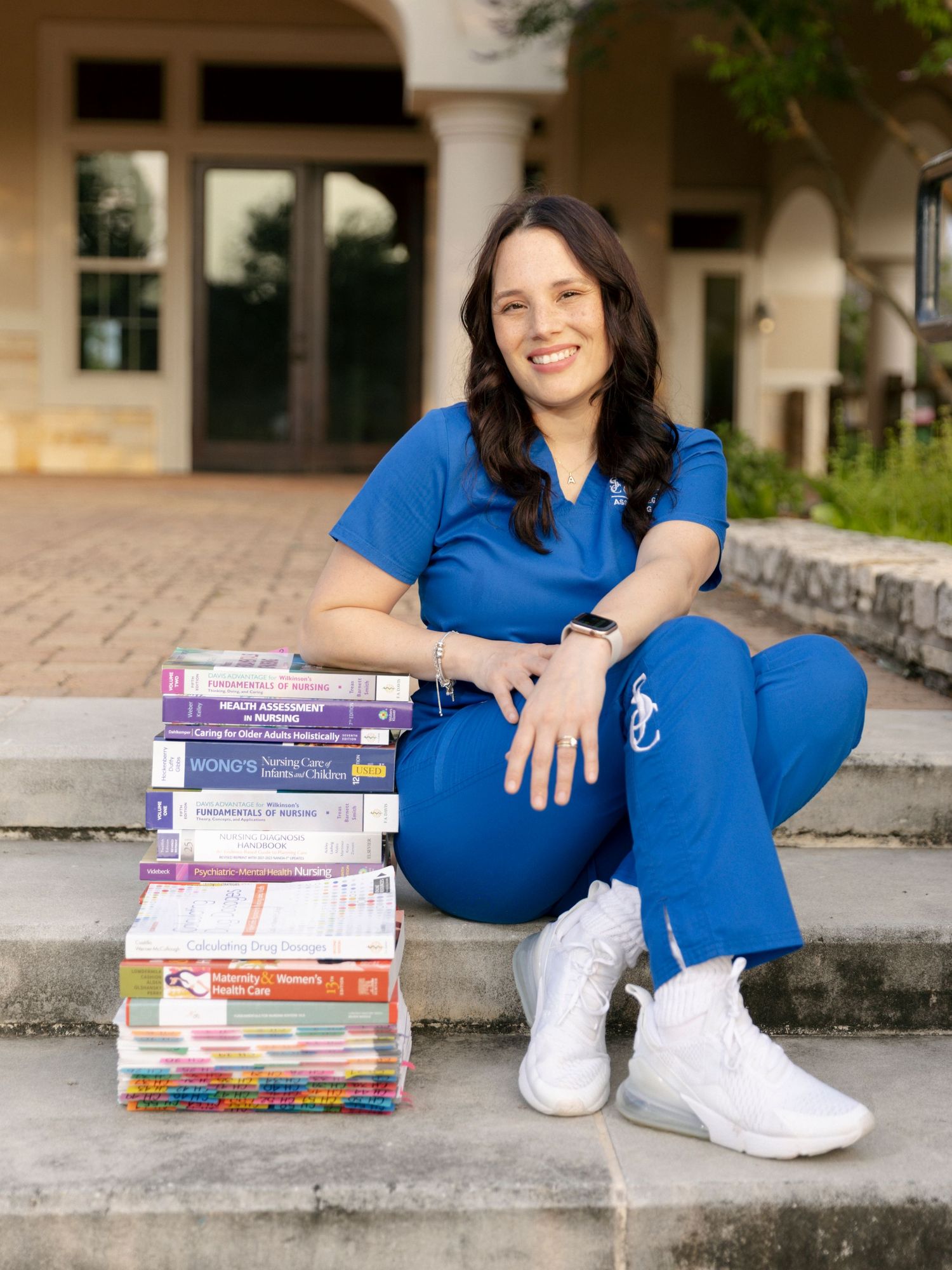 A healthcare worker in blue scrubs poses with a stack of nursing textbooks on outdoor concrete steps.