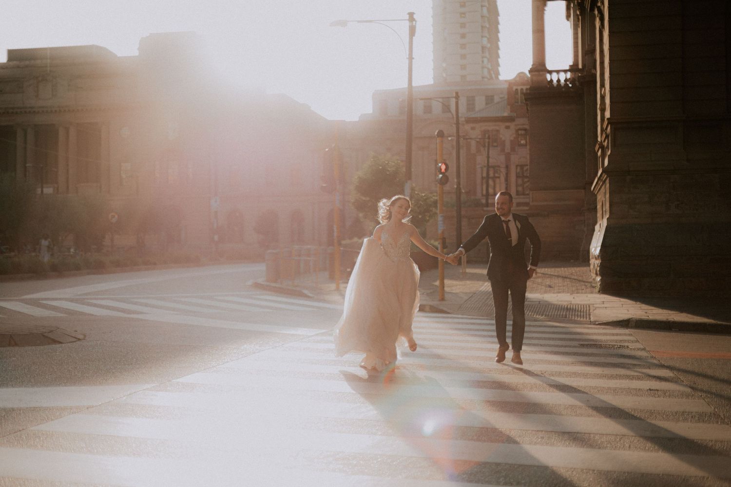 A couple walks hand in hand down a sunlit city street with tall buildings in the background.