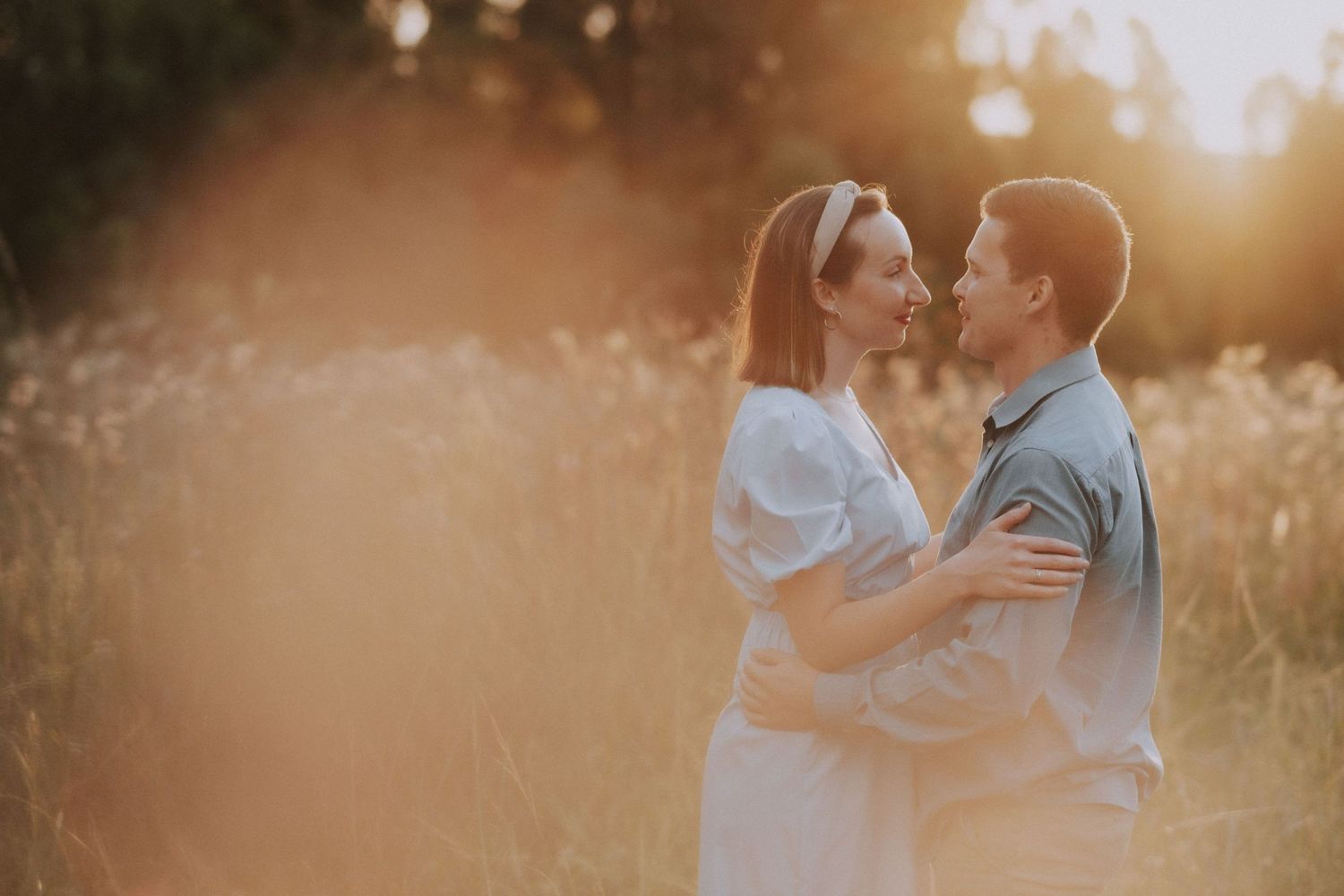 A couple in light colored clothing sharing an intimate moment in a sun-lit field.