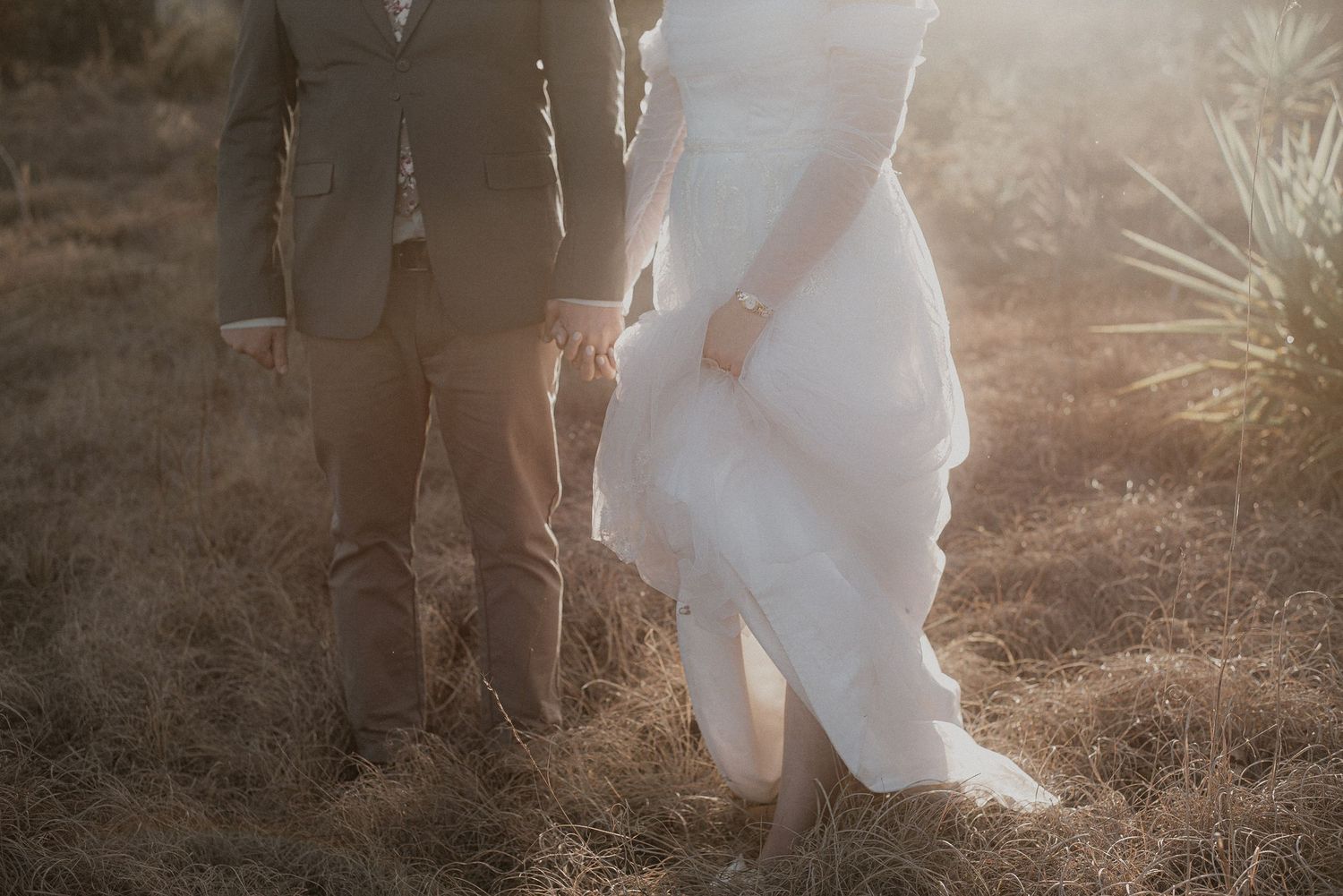 A couple in wedding attire stands together in golden sunlight against a natural backdrop.