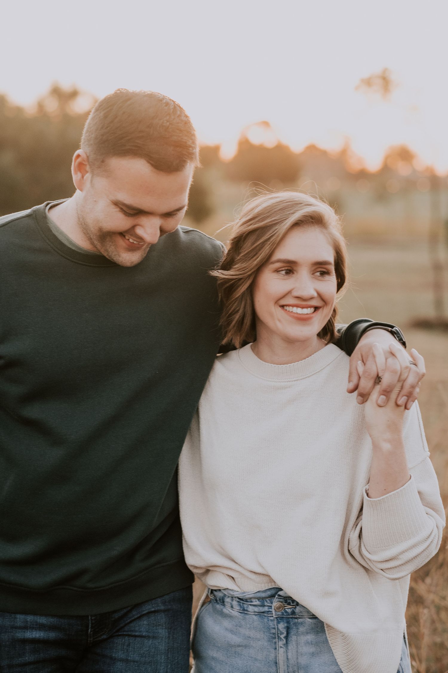 A romantic couple wearing casual sweaters embrace and smile during a golden hour photoshoot outdoors.