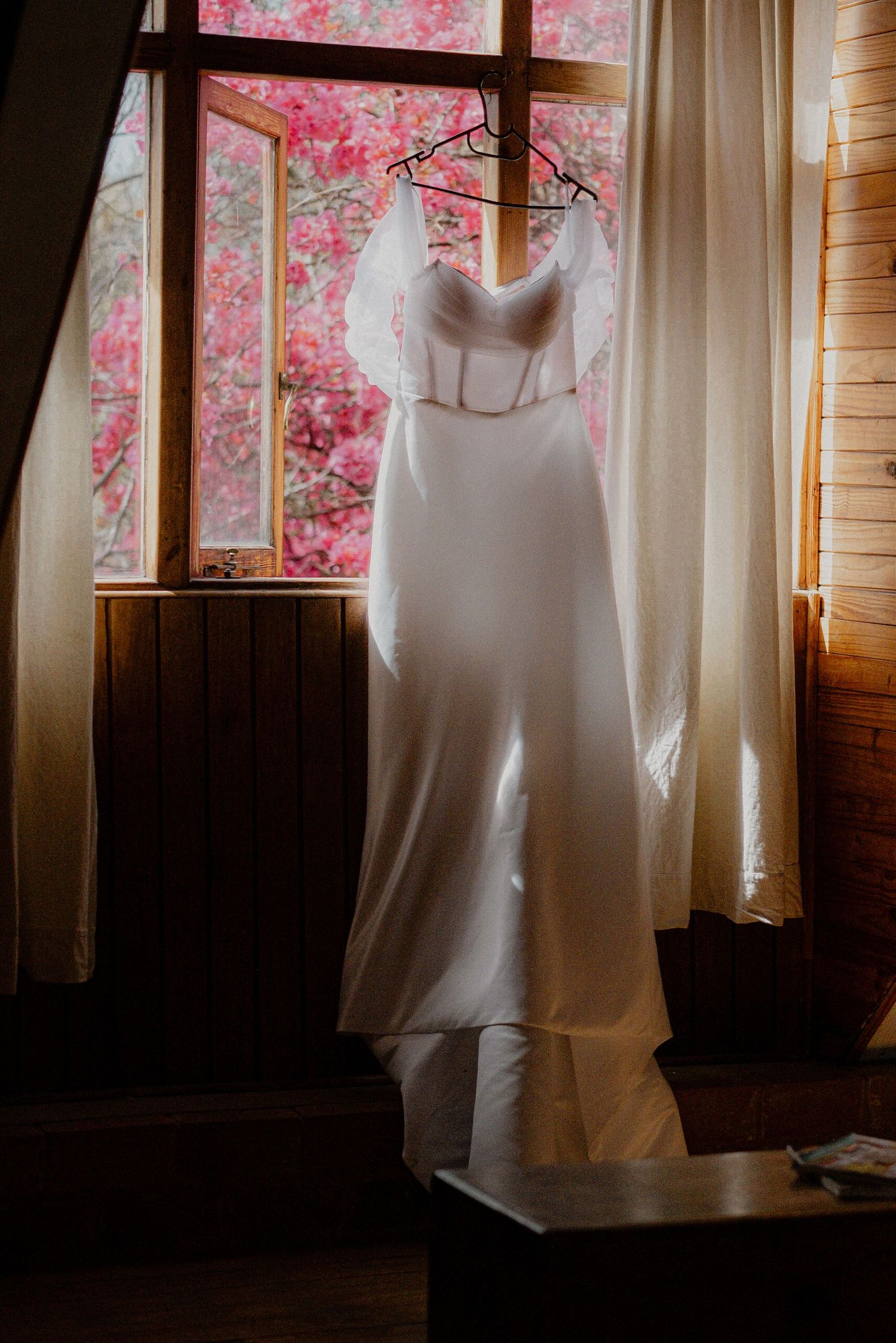 A white wedding dress hangs in front of a window with pink cherry blossoms visible outside.